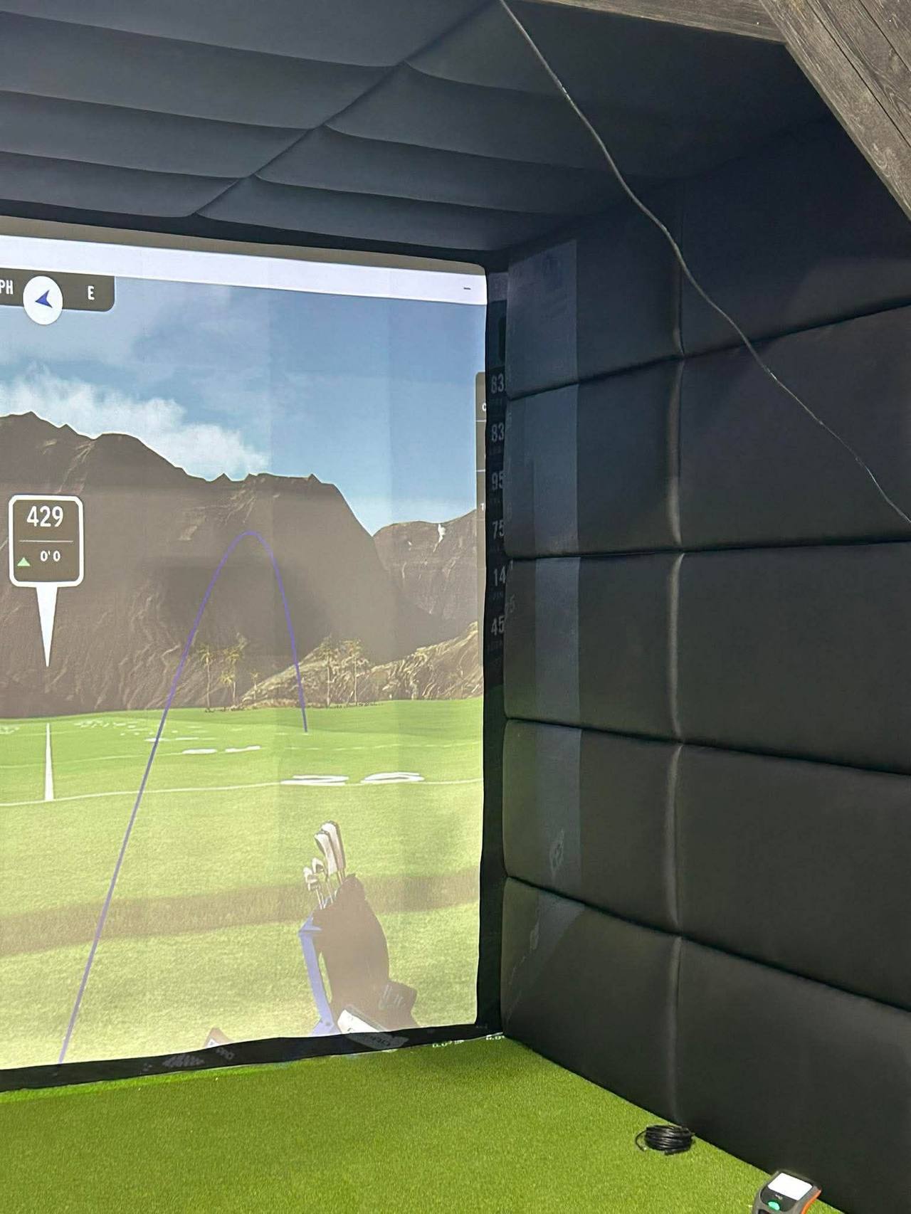 Golf simulator bay from side angle showing black tufted wall panels on side wall and ceiling, screen showing tropical golf course