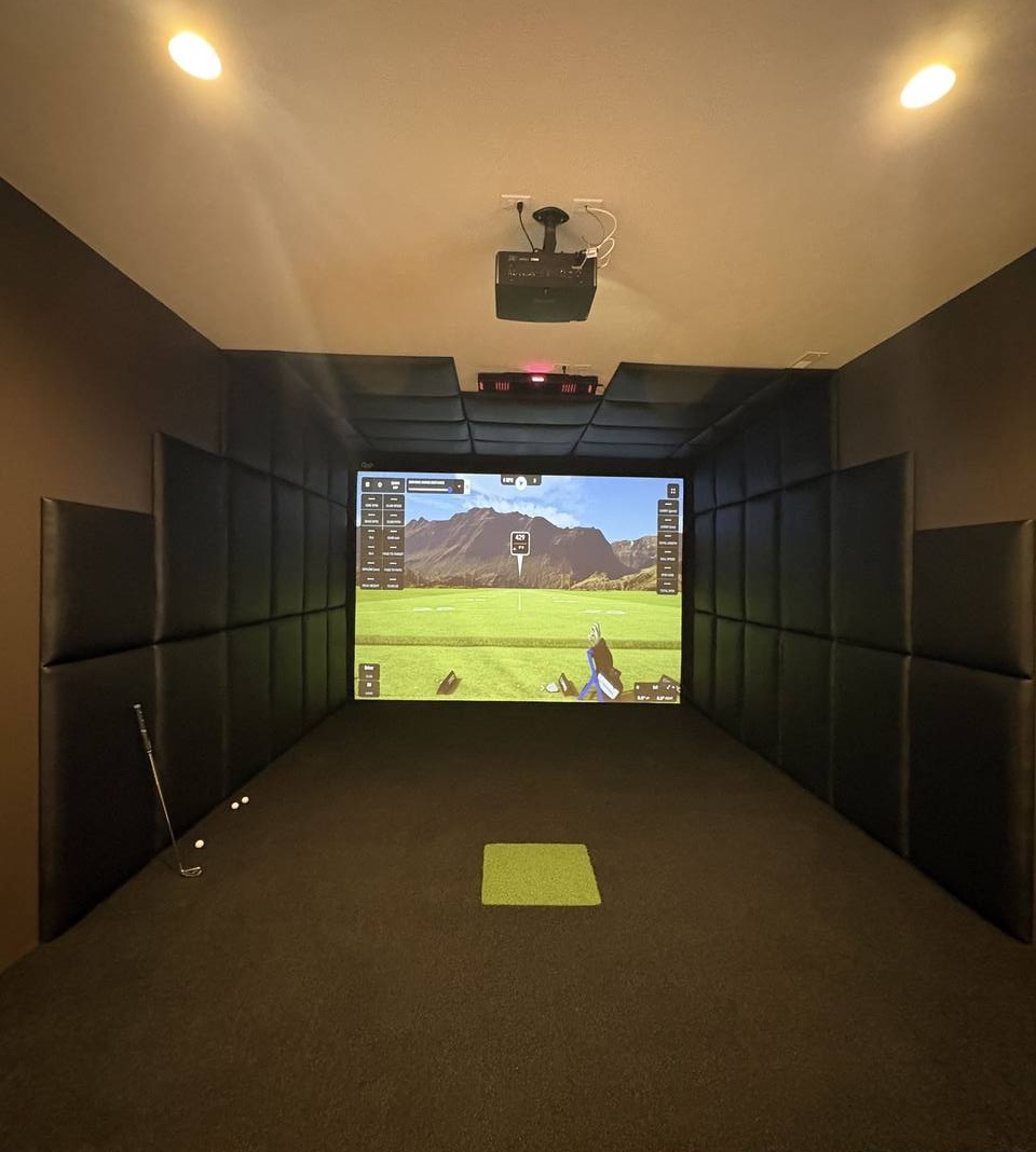 Real indoor golf simulator room with black tufted Shank Pro wall panels on both side walls and around impact screen, dark carpet floor with green turf hitting mat